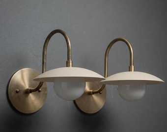 May include: Two wall-mounted sconces with a mid-century modern design. Each sconce features a circular brass base, a curved brass arm, and a cream-colored, saucer-shaped shade. The shades encase a white, globe-shaped light fixture.