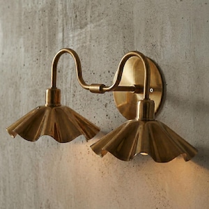 May include: A brass wall sconce with two lights. Each light has a ruffled shade and a curved arm. The sconce is mounted on a round backplate. The fixture is made of brass and is mounted on a concrete wall.