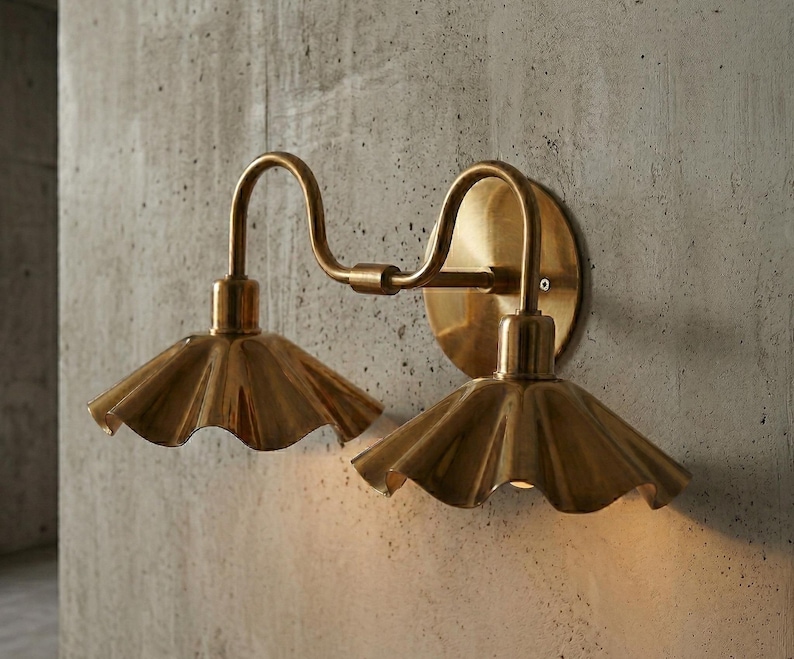 May include: A brass wall sconce with two lights. Each light has a ruffled shade and a curved arm. The sconce is mounted on a round backplate. The fixture is made of brass and is mounted on a concrete wall.