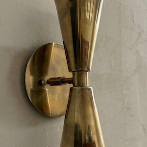 Op de afbeelding: Een messing wandlamp met een conisch kapontwerp. De lamp heeft een cirkelvormige basis en een draaiarm die de kap met de basis verbindt. De afwerking is warm, gepolijst messing, dat het licht reflecteert.