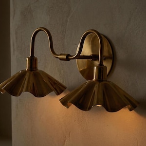 May include: A brass wall sconce with two lights. Each light has a ruffled shade and a curved arm. The sconce is mounted on a round backplate. The fixture is made of brass and has a warm, golden finish.