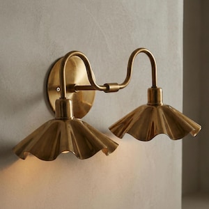 May include: A brass wall sconce with two lights. The sconce has a circular backplate and curved arms. Each light has a ruffled shade. The fixture is made of brass and is mounted on a light-colored wall.