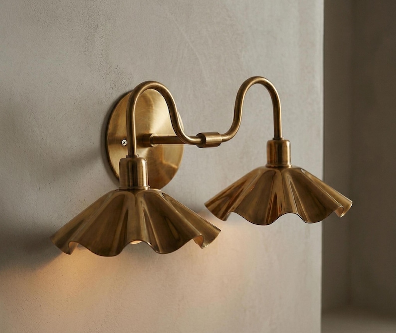 May include: A brass wall sconce with two lights. The sconce has a circular backplate and curved arms. Each light has a ruffled shade. The fixture is made of brass and is mounted on a light-colored wall.