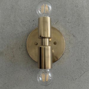 May include: A brass wall sconce featuring two clear, round light bulbs. The fixture has a circular backplate and cylindrical elements. The bulbs have visible filaments, mounted on a grey concrete wall.