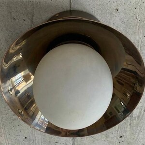 May include: A modern ceiling light fixture with a bronze-colored, bowl-shaped shade and a large, white globe light. The fixture is mounted on a concrete ceiling. The light reflects the surrounding environment.