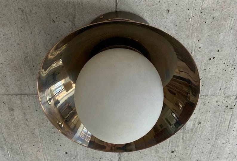 May include: A modern ceiling light fixture with a bronze-colored, bowl-shaped shade and a large, white globe light. The fixture is mounted on a concrete ceiling. The light reflects the surrounding environment.