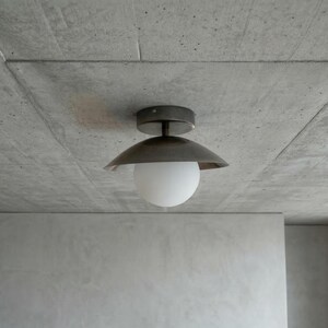 May include: A modern ceiling light fixture with a dark metal shade and a white globe. The light is mounted on a gray concrete ceiling. The room has a minimalist design.