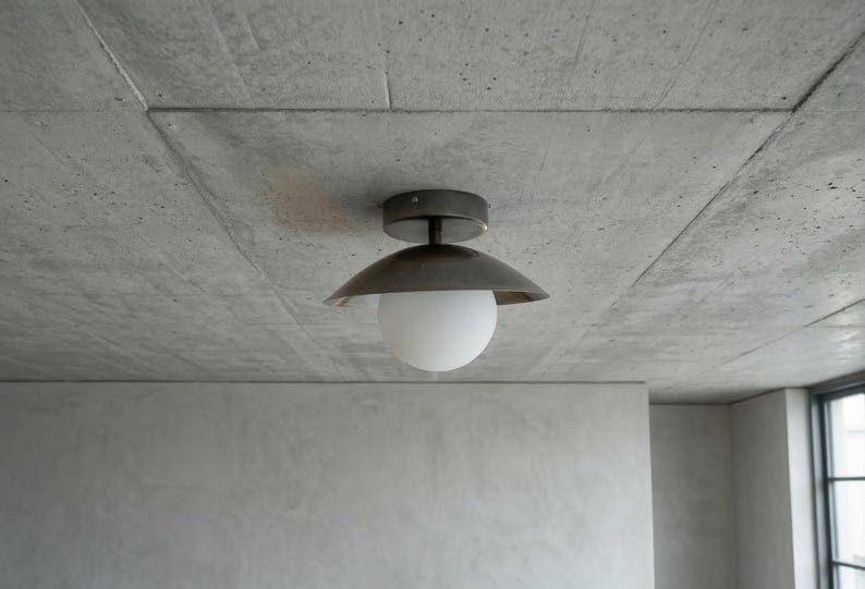 May include: A modern ceiling light fixture with a dark metal shade and a white globe. The light is mounted on a gray concrete ceiling. The room has a minimalist design.