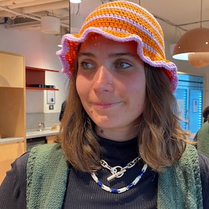 Peut inclure: Un chapeau crocheté à la main avec un bord ondulé. Le chapeau présente des rayures alternées orange et violet clair, avec une bordure rose clair. Un accessoire élégant pour se protéger du soleil ou pour la mode.