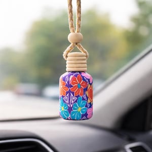 May include: A decorative car air freshener with a floral design, suspended from a car's rearview mirror. The bottle has a wooden top and is adorned with a vibrant floral pattern in purple, red, blue, and pink. The hanging cord is a beige, braided rope.