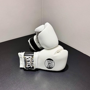 Peut inclure: Une paire de gants de boxe blancs avec des sangles de poignet noires. Les gants présentent le logo Cleto Reyes en noir et blanc sur le poignet et le dos de la main. Le texte sur la sangle indique Cleto Reyes Hecho en Mexico Profesional.