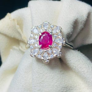 May include: A silver ring with a central oval ruby, encircled by pear-shaped white gemstones. The ruby is a vibrant red, and the ring's design is floral. The gemstones are arranged to resemble petals, creating a striking visual.