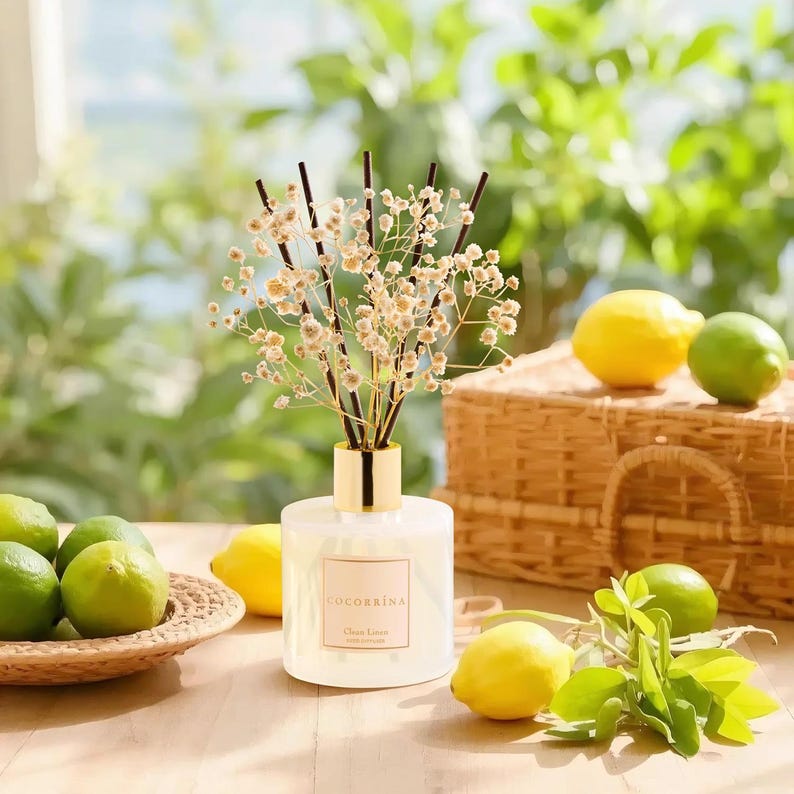 Pu&ograve; includere: Un diffusore a bastoncini in vetro trasparente con un collare color oro, contenente un liquido chiaro e sormontato da bastoncini scuri e fiori secchi bianchi di gipsofila. L'etichetta sul davanti riporta la scritta COCORRINA Clean Linen.