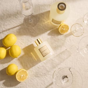 Pu&ograve; includere: Una bottiglia di vetro quadrata con tappo dorato etichettata COCORRINE, una grande bottiglia rotonda con tappo scuro, quattro limoni gialli interi, mezzo limone e tre calici di vetro vuoti disposti su una superficie in tessuto beige.