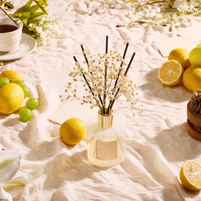 Pu&ograve; includere: Un diffusore di fragranze in vetro con collare dorato, contenente liquido trasparente e bastoncini neri sormontati da fiori secchi di gipsofila. La bottiglia presenta un'etichetta quadrata dorata ed &egrave; circondata da limoni gialli e uva verde.