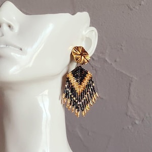 May include: Gold-toned earrings with a geometric design. The earrings feature a circular stud, a black and gold beaded central section, and beaded fringe. The earrings are displayed on a white mannequin head against a gray background.