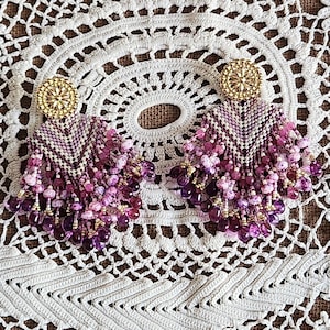Può includere: Un paio di orecchini fatti a mano con un perno circolare color oro. Gli orecchini presentano un design di perline nelle tonalità del viola, del rosa e del bianco, con perline pendenti di varie dimensioni e tonalità. Gli orecchini sono esposti su un centrino di pizzo bianco.