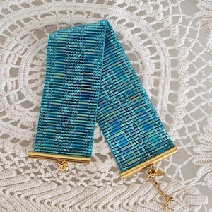 May include: A woven bracelet in shades of teal, turquoise, and blue. The bracelet features a rectangular design with gold-colored metal end caps and a delicate gold chain. The bracelet is displayed on a white, lacy surface.