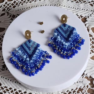 Può includere: Un paio di orecchini fatti a mano con perline nei toni del blu, a forma di ventaglio. Gli orecchini presentano un motivo geometrico e perline pendenti. In cima ci sono borchie circolari color oro. Gli orecchini sono esposti su una superficie bianca.