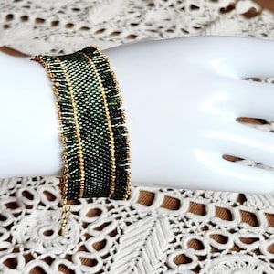 Peut inclure: Un bracelet de perles avec un motif à carreaux vert et noir et des accents dorés. Le bracelet est porté sur une main de mannequin blanche. Le bracelet est fait de petites perles et a un design délicat et complexe.