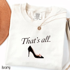 Peut inclure: Un t-shirt à manches courtes ivoire avec un col rond et une étiquette de marque Comfort Colors. Le t-shirt présente le texte marron foncé That's all. imprimé au-dessus d'un graphique d'escarpin noir à talon avec un petit nœud rose clair.