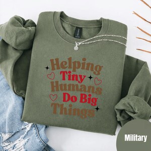 Peut inclure: Sweat-shirt vert olive avec l'inscription "Helping Tiny Humans Do Big Things" en marron et rouge. Le sweat-shirt a un col rond et des manches longues. Un collier argenté avec un pendentif est sur le sweat-shirt.