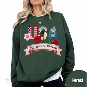 Peut inclure: Sweat-shirt vert forêt avec le mot "USA" aux couleurs patriotiques et des images. Le "U" est un drapeau américain, le "S" représente un aigle et le "A" montre la Statue de la Liberté. Une bannière indique "250 years OF freedom".