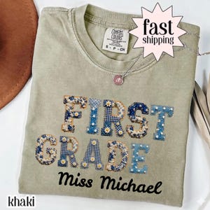 Peut inclure: Un t-shirt à col rond kaki avec le texte First Grade en grandes lettres de style bloc remplies de motifs floraux et vichy bleus, agrémentées de petites marguerites blanches. En dessous, le nom Miss Michael est imprimé en écriture cursive noire.