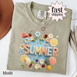 Może przedstawiać: T-shirt w kolorze khaki z nadrukiem graficznym słowa SUMMER wypukłymi, wielokolorowymi literami. Wzór otoczony jest ilustracjami truskawek, kawałków arbuza, stokrotek, plasterków owoców cytrusowych oraz ananasa.
