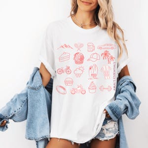 Peut inclure: Un t-shirt blanc orné d'une grille d'icônes en ligne rouge représentant des montagnes, une raquette, une chope de bière, une voiture, un gâteau, un panier, une casquette, des palmiers, un vélo, la météo, des planches, des skis, un burger, une moto, un cupcake et un haltère, avec le texte AMAL'S ESSENTIALS imprimé verticalement.