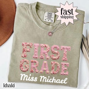 Peut inclure: Un t-shirt à manches courtes de couleur kaki avec le texte FIRST GRADE en lettres appliquées à motifs roses ornées de petites fleurs blanches et de perles, suivi du texte cursif blanc Miss Michael et un collier argenté.