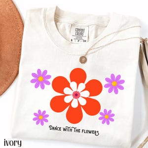 Peut inclure: Un t-shirt à manches courtes ivoire orné d'un grand motif floral central aux pétales orange et blancs, entouré de quatre petites fleurs violettes. Le texte "DANCE WITH THE FLOWERS" est imprimé sous le motif dans une police serif noire.