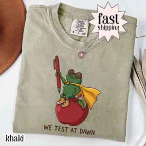 Peut inclure: Un t-shirt kaki avec une illustration représentant une grenouille verte portant une cape jaune, des lunettes rouges et des baskets jaunes, assise sur une pomme rouge tout en tenant un stylo rouge. Le texte "WE TEST AT DAWN" est imprimé en dessous.