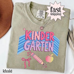 Peut inclure: Un t-shirt kaki avec le texte KINDERGARTEN en lettres roses et violettes sur un motif d'arc-en-ciel à rayures bleues. Sous le texte se trouvent un crayon rose, un nœud rose et une pomme en dégradé. Le t-shirt porte une étiquette Comfort Colors.