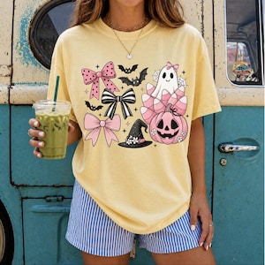 Peut inclure: Un t-shirt à manches courtes jaune pâle avec une impression graphique composée de nœuds roses et noirs, de chauves-souris, d'un fantôme blanc avec un nœud rose, d'une citrouille rose et d'un chapeau de sorcière noir à fleurs blanches.