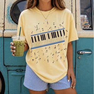 Peut inclure: Un t-shirt à manches courtes jaune pâle avec une impression graphique d'une section de clavier de piano encadrée par deux lignes bleues horizontales, entourée de notes de musique noires éparpillées. Porté avec un short à rayures bleu et blanc.