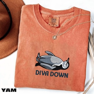 May include: A coral-colored t-shirt with a crew neck and short sleeves. The shirt features a graphic of a stylized penguin with a decorative pattern and the text "DIVA DOWN" in dark blue. A gold necklace with a pendant is also visible.
