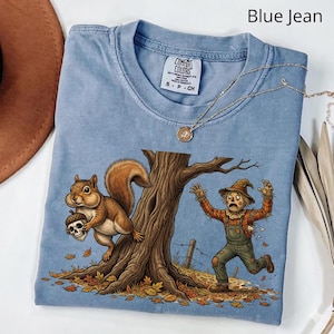 Può includere: Una t-shirt color blu jeans con una stampa grafica raffigurante uno scoiattolo che tiene una ghianda con motivo a teschio e uno spaventapasseri spaventato che scappa vicino a un albero. La maglietta ha un girocollo e un'etichetta Comfort Colors.