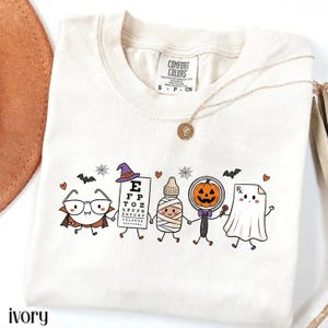 May include: An ivory t-shirt featuring a graphic of five cartoon characters holding hands: a vampire with glasses, an eye chart wearing a witch hat, a mummy, a pumpkin with a magnifying glass, and a ghost holding a prescription paper.