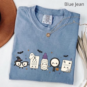 Könnte beinhalten: Ein blaues T-Shirt mit einer Grafik von fünf niedlichen, anthropomorphen Charakteren, die sich an den Händen halten: eine Brille mit Hexenhut, ein Sehtest-Diagramm, eine Mumie, eine Lupe mit Kürbis und ein Rezeptblock mit einem Spinnennetz.