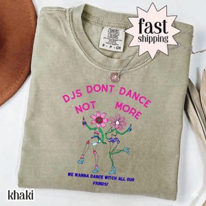 Può includere: T-shirt color cachi con la scritta rosa "DJs Don't Dance Not More". La maglietta presenta una grafica di due figure con la testa a forma di fiore che tengono bottiglie. Il testo "We Wanna Dance Witch All Our Frinds!" è sotto.