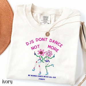 Peut inclure: T-shirt Comfort Colors ivoire avec l'inscription rose "DJs Don't Dance Not More". Le t-shirt présente un motif de deux personnages dansant avec des têtes de fleurs. Un collier en or est drapé sur le t-shirt.