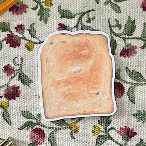 May include: A sticker of a slice of toasted bread with a white border. The toast is light brown with darker brown areas, suggesting a toasted texture. The sticker is on a floral patterned fabric with a pencil, scissors, and a gold paper clip.