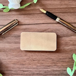 Op de afbeelding: Een goudkleurige vulpen met een zwarte grip en een gouden penpunt, een bijpassende penkap en een blanco rechthoekige kaart liggen op een houten oppervlak. Versierd met witte papieren bloemen en groene papieren bladeren.