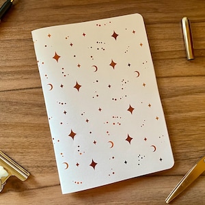Op de afbeelding: Een wit notitieboek met een herhalend patroon van koperkleurige sterren, maansikkels en kleine stippen. Het notitieboek is omringd door een goudkleurige pen, een goudkleurige metalen binderclip, een penkapje en een schaar met gouden handvatten.
