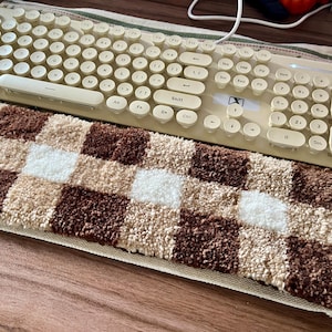 Peut inclure: Un clavier mécanique crème avec des touches rondes est posé au-dessus d'un tapis à carreaux marron, beige et blanc. Le clavier a un design rétro. Le tapis est rectangulaire et a un aspect doux et texturé.