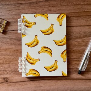 May include: A spiral-bound notebook with a white cover featuring a repeating pattern of yellow bananas. To the left of the notebook is a gold-toned metal binder clip, and to the right is a clear and black ballpoint pen with a pocket clip.
