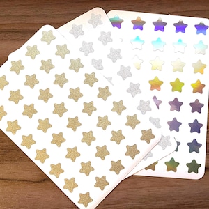 Puede incluir: Hojas de pegatinas con forma de estrella en purpurina dorada, purpurina plateada y acabados iridiscentes. Las pegatinas están dispuestas en filas sobre hojas de respaldo blancas. El fondo es una superficie de madera oscura.
