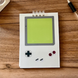 May include: A white notepad designed to resemble a retro handheld gaming device. It features a green screen, gray accents, and the classic button layout with the words "SELECT" and "START". A pen rests nearby.
