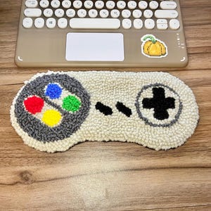 May include: A white and gray needlepoint rug shaped like a classic video game controller. The controller has four colored buttons, a directional pad, and a black plus button.